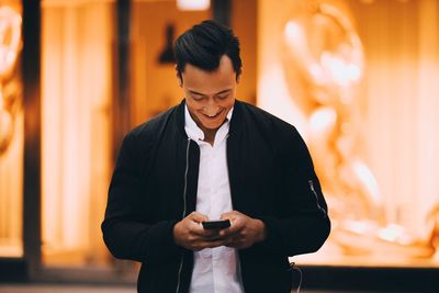 Smiling young man using mobile phone while standing in illuminated city