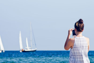 Rear view of woman photographing of boat in sea through mobile phone on sunny day