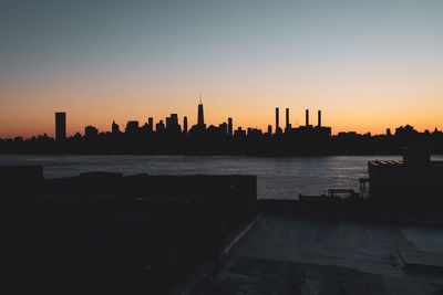 Silhouette buildings by river against sky during sunset