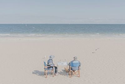 Rear view of people sitting in chairs on beach against sky