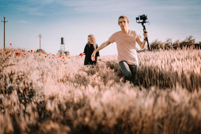 Young woman and man running on field with camera in hand against sky during summer