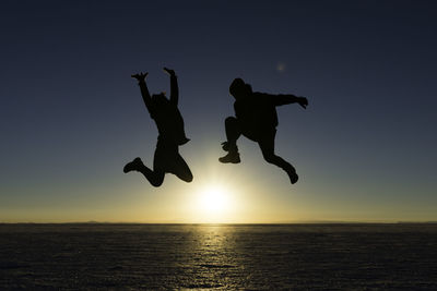 Silhouette people jumping on sea against sky during sunset