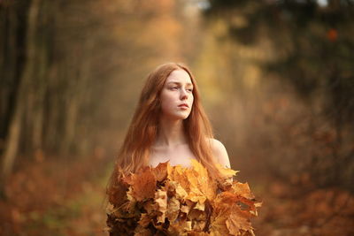 Thoughtful young woman covered with leaves standing at forest during autumn