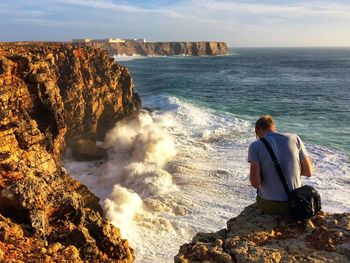 Rear view of man sitting on cliff by sea