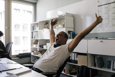 Happy male entrepreneur screaming with arms raised while sitting at office