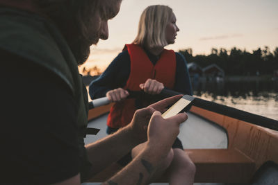 Man using mobile phone while woman rowing boat on lake