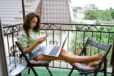 Young woman sits on a balcony at a table working with a phone behind a laptop