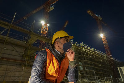 Occupational safety. construction worker posing at night wearing safety vest, hardhat, medical mask