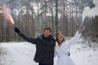 Smiling wedding couple holding firework and distress flare while standing at forest during winter