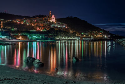 Illuminated buildings by lake against sky at night