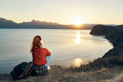 Woman sitting by lake against sky during sunset