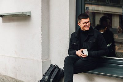 Smiling young man looking away while sitting on window sill