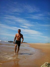 Shirtless man with headphones walking on beach against cloudy sky on sunny day