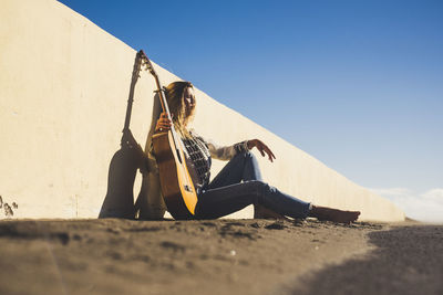 Young woman with guitar sitting on road by wall against clear sky