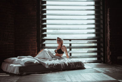 Portrait of young woman wearing bikini sitting on bed against window at home