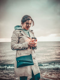 Woman holding illuminated jar while standing at beach against sky