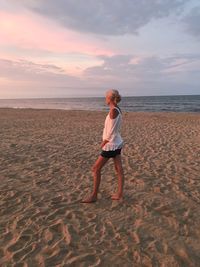 Full length side view of woman looking at sea while standing on sand against sky during sunset