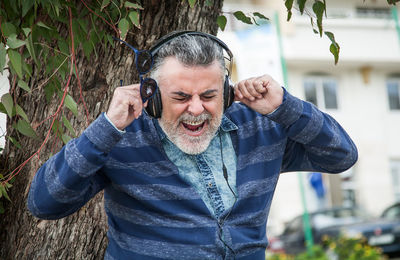 Mature man screaming while listening music against tree trunk in park
