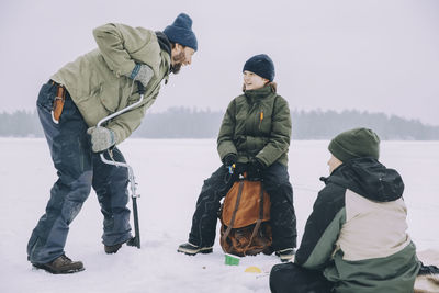 Happy mature man using ice auger while talking with son sitting on backpack at frozen lake