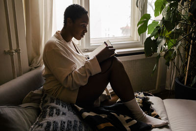 Side view of young woman writing in book while sitting on bed at home