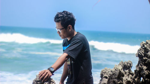 Young man standing on rock by sea against sky