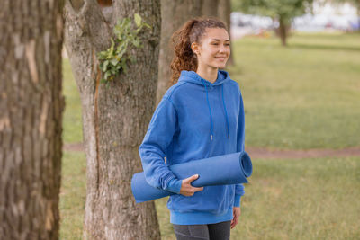 Smiling young woman holding exercise mat at park