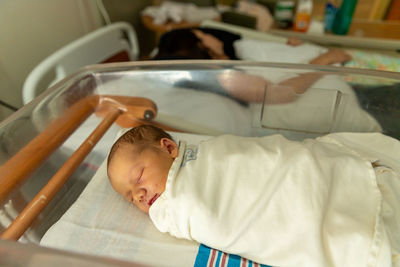 Newborn baby in swaddle sleeping in hospital room near mother.