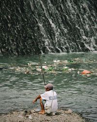 Rear view of man sitting on rock by river
