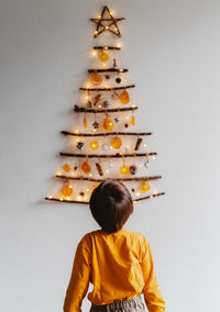 Rear view of child standing in front of handmade craft christmas tree hanging on wall. 