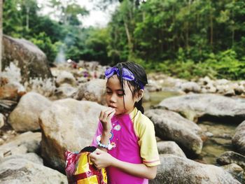Girl eating food while standing at riverbank in forest