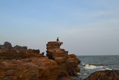 Low angle view of man sitting on cliff by sea against sky