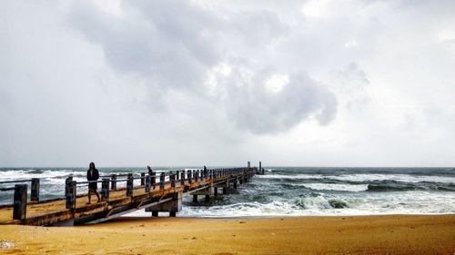 Man walking on pier at beach against cloudy sky