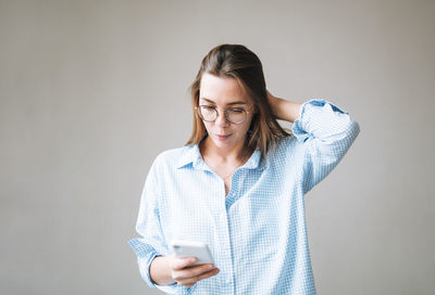 Portrait of young smiling woman with dark long hair in blue shirt using mobile phone in hand on grey 