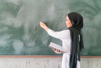 Side view of teacher wearing traditional clothing writing on blackboard while standing in classroom