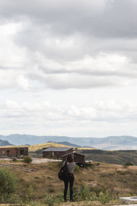 Rear view of woman standing on rock while looking at houses on mountain against sky