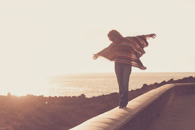 Woman balancing on retaining wall by sea against clear sky during sunny day