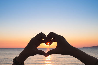 Hand holding heart shape against sea during sunset