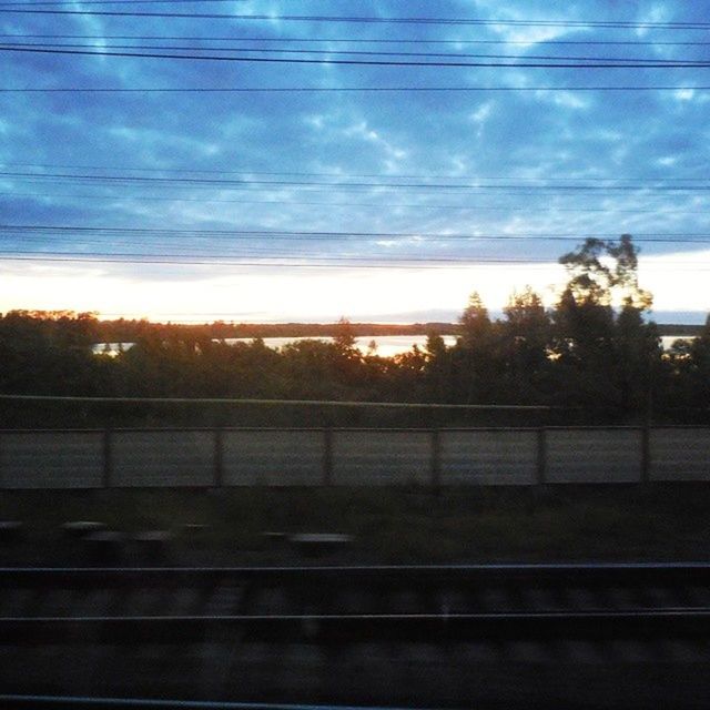 railroad track, rail transportation, transportation, sky, power line, public transportation, cloud - sky, electricity pylon, tree, sunset, railroad station, train - vehicle, railway track, cable, railroad station platform, power supply, electricity, cloud, train, connection