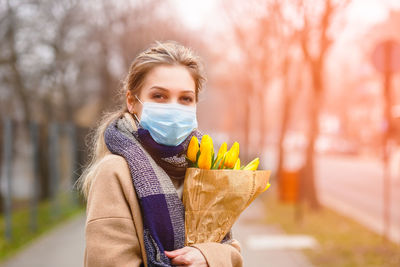 A young girl in a beige coat with blonde hair is holding a bouquet of yellow tulips, wearing a mask.