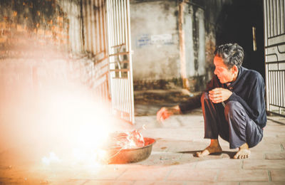 Mature man with blowing fire in container while crouching outdoors at night