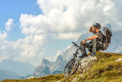 Man sitting with bicycle on mountain against cloudy sky
