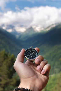 A hand with a compass against the backdrop of epic snow-capped mountains with clouds and a forest at