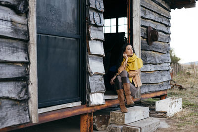 Stylish woman in boots and scarf sitting on porch of old wooden house in countryside looking away