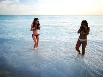 Happy female friends standing in shallow water at beach against sky