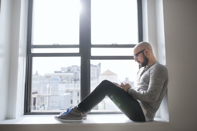 Side view of man using smart phone while sitting on window