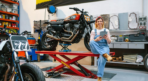 Smiling mechanic woman looking at camera while holding digital tablet sitting over platform