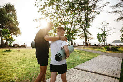 Father with arm around son carrying exercise mats walking in park during sunset
