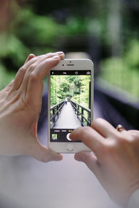 Cropped image of woman photographing person on footbridge through smart phone