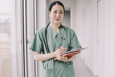 Portrait of confident female physician holding clipboard while standing in hospital corridor