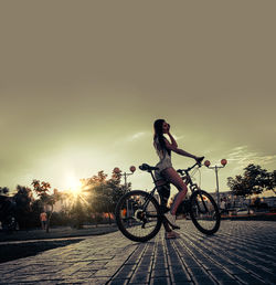 Woman riding bicycle on street against sky during sunset